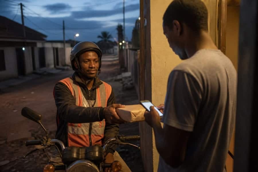 Motorcycle courier handing a parcel to a customer checking a phone at the door